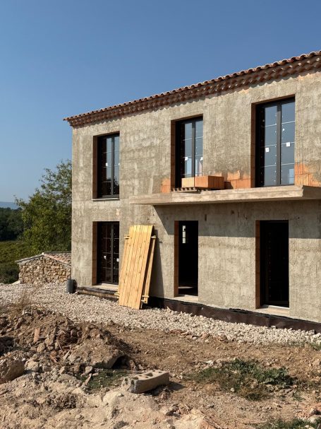 Construction d'une maison avec fenêtres, balcon et extérieur en béton. Terrain en cours d'aménagement.